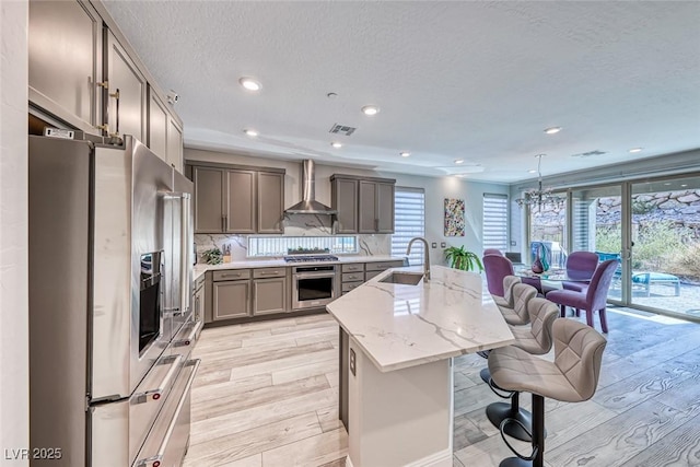 kitchen with pendant lighting, sink, appliances with stainless steel finishes, a kitchen island with sink, and wall chimney exhaust hood