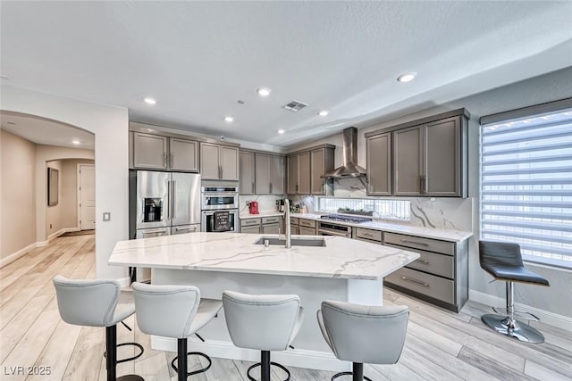 kitchen featuring appliances with stainless steel finishes, an island with sink, sink, light stone counters, and wall chimney range hood