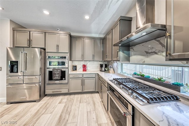 kitchen with gray cabinetry, tasteful backsplash, stainless steel appliances, light stone countertops, and wall chimney range hood