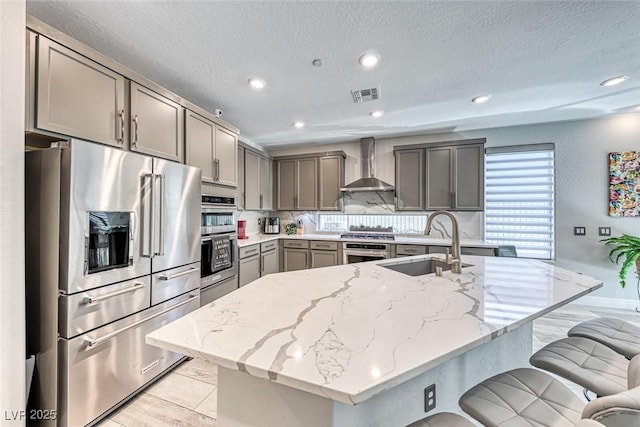 kitchen featuring sink, gray cabinetry, light stone counters, stainless steel appliances, and wall chimney exhaust hood