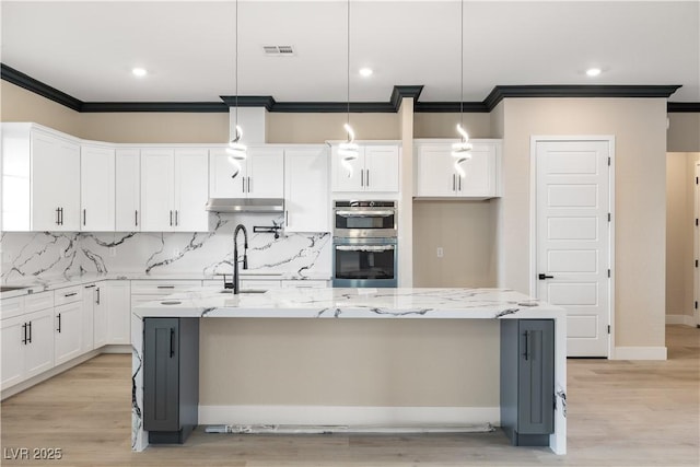 kitchen featuring double oven, decorative light fixtures, an island with sink, white cabinets, and light stone countertops