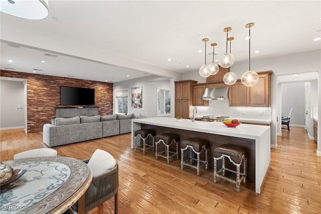 kitchen with pendant lighting, a breakfast bar area, a center island with sink, light wood-type flooring, and wall chimney exhaust hood