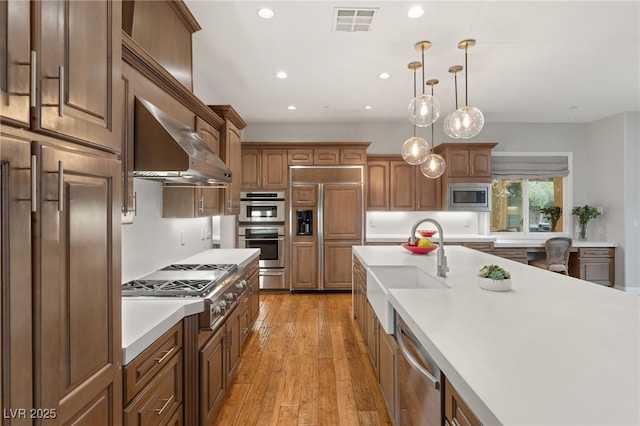 kitchen with sink, light hardwood / wood-style flooring, built in appliances, decorative light fixtures, and wall chimney exhaust hood