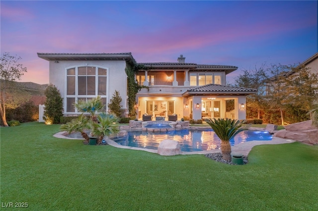 back house at dusk with a swimming pool with hot tub, a balcony, and a lawn