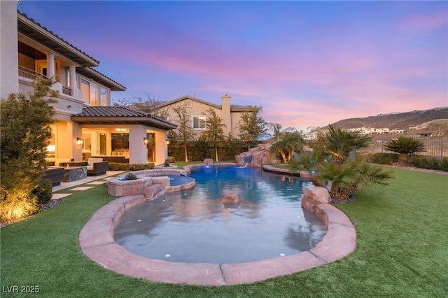 pool at dusk with a water slide, an outdoor living space, a lawn, and an in ground hot tub