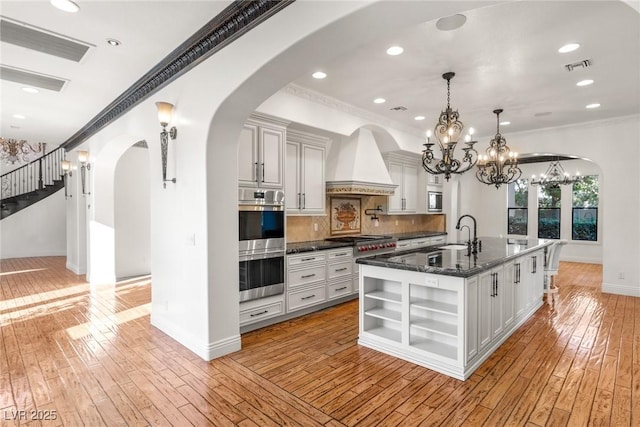 kitchen featuring a kitchen island with sink, stainless steel appliances, ornamental molding, custom exhaust hood, and dark stone counters