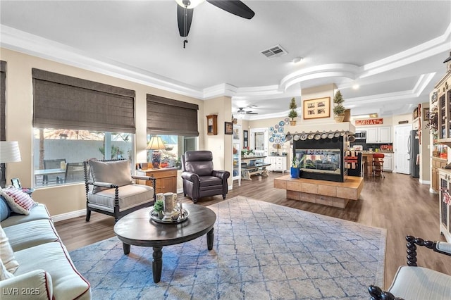 living room featuring a tile fireplace, ceiling fan, a tray ceiling, wood-type flooring, and ornamental molding