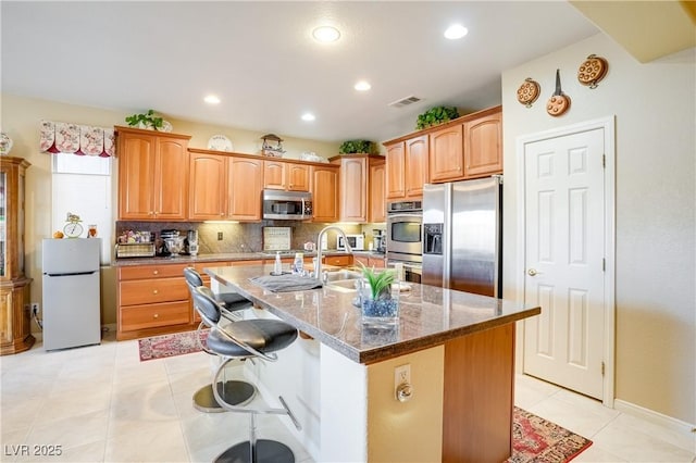 kitchen with visible vents, appliances with stainless steel finishes, a kitchen island with sink, dark stone counters, and a kitchen bar