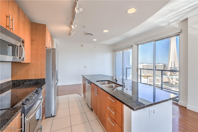 kitchen featuring sink, dark stone countertops, stainless steel appliances, track lighting, and a center island with sink