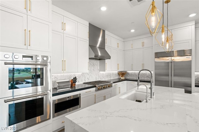 kitchen with decorative light fixtures, white cabinetry, sink, stainless steel appliances, and wall chimney range hood