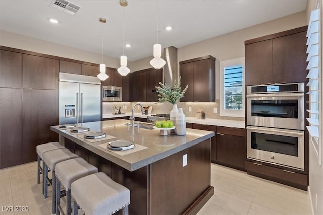 kitchen with sink, a kitchen island with sink, built in appliances, dark brown cabinetry, and decorative light fixtures