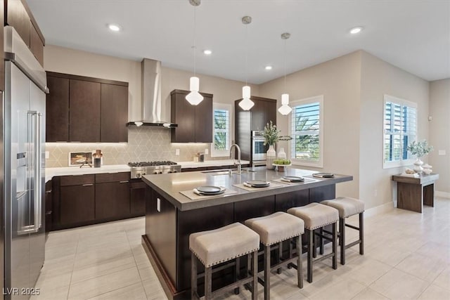 kitchen with sink, wall chimney range hood, hanging light fixtures, stainless steel appliances, and a center island with sink