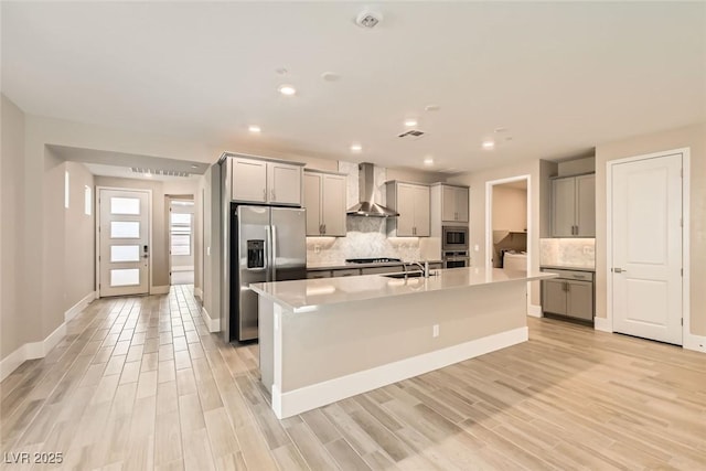 kitchen with an island with sink, wall chimney range hood, appliances with stainless steel finishes, and gray cabinetry