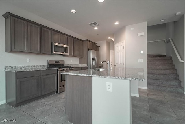 kitchen with sink, dark brown cabinets, stainless steel appliances, light stone countertops, and a center island with sink