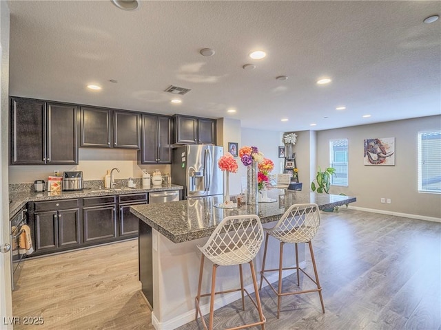 kitchen featuring an island with sink, a kitchen bar, dark stone counters, light hardwood / wood-style floors, and stainless steel appliances