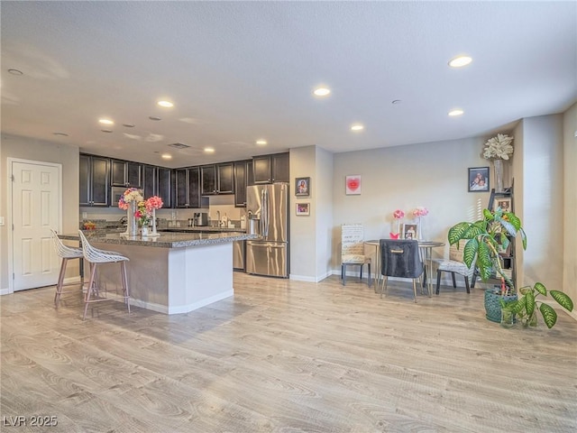 kitchen featuring a breakfast bar area, stainless steel refrigerator with ice dispenser, light hardwood / wood-style floors, a center island with sink, and dark stone counters