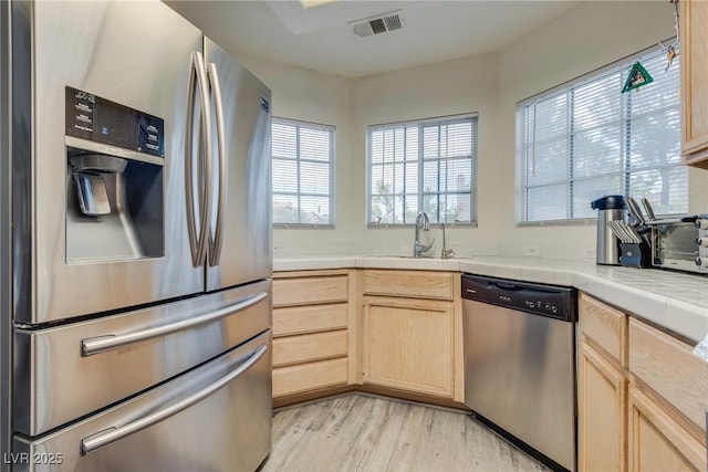 kitchen featuring appliances with stainless steel finishes, tile countertops, light brown cabinetry, sink, and light hardwood / wood-style flooring