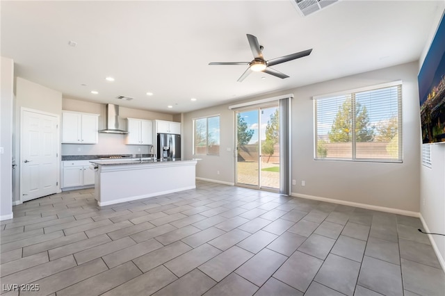 kitchen featuring stainless steel refrigerator with ice dispenser, white cabinetry, ceiling fan, a kitchen island with sink, and wall chimney range hood