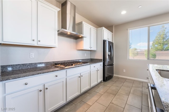 kitchen featuring wall chimney range hood, light tile patterned floors, stainless steel appliances, white cabinets, and dark stone counters