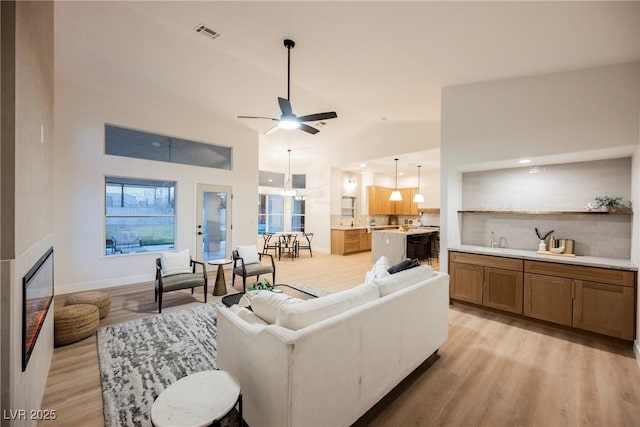 living room with high vaulted ceiling, a fireplace, sink, ceiling fan, and light hardwood / wood-style flooring