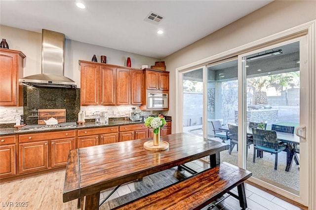 kitchen featuring appliances with stainless steel finishes, light wood-type flooring, dark stone counters, wall chimney exhaust hood, and tasteful backsplash