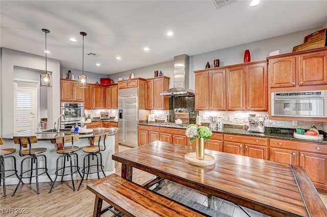 kitchen with stainless steel appliances, hanging light fixtures, dark stone countertops, wall chimney range hood, and decorative backsplash