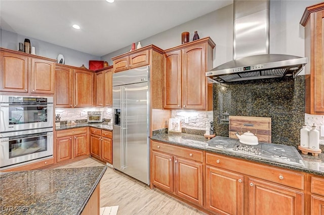 kitchen with dark stone counters, stainless steel appliances, wall chimney range hood, and backsplash