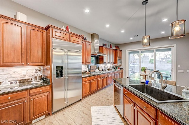 kitchen with hanging light fixtures, stainless steel appliances, sink, dark stone counters, and wall chimney exhaust hood