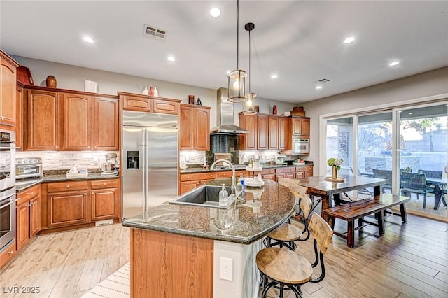 kitchen with appliances with stainless steel finishes, wall chimney exhaust hood, an island with sink, pendant lighting, and sink