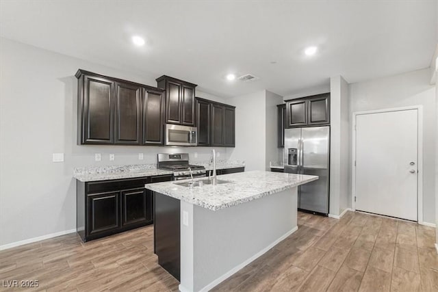 kitchen featuring sink, a center island with sink, stainless steel appliances, light stone countertops, and light hardwood / wood-style floors