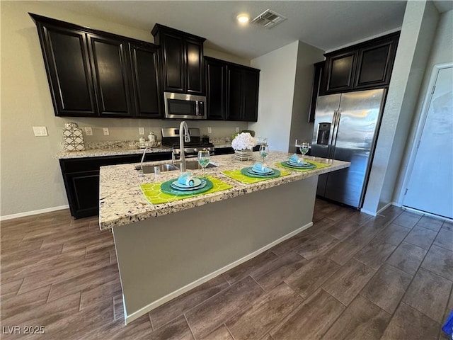 kitchen with stainless steel appliances, an island with sink, sink, and light stone counters