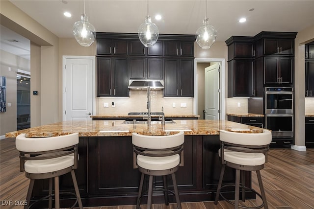 kitchen with light stone counters, dark wood-style flooring, stainless steel double oven, under cabinet range hood, and a sink