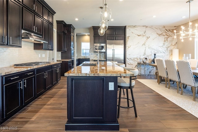 kitchen featuring light stone counters, a kitchen breakfast bar, built in appliances, under cabinet range hood, and a sink