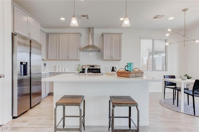 kitchen with an island with sink, appliances with stainless steel finishes, pendant lighting, and wall chimney exhaust hood