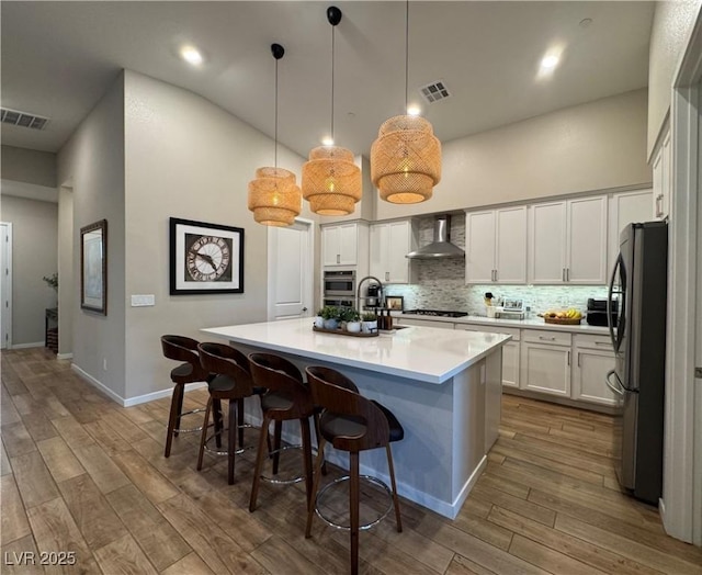 kitchen with pendant lighting, an island with sink, white cabinets, stainless steel fridge, and wall chimney range hood