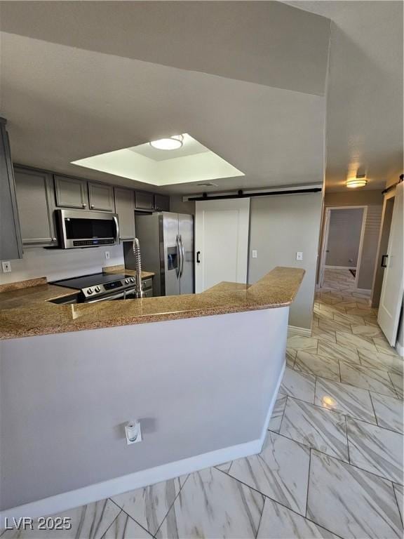 kitchen with appliances with stainless steel finishes, kitchen peninsula, gray cabinets, a barn door, and light stone countertops