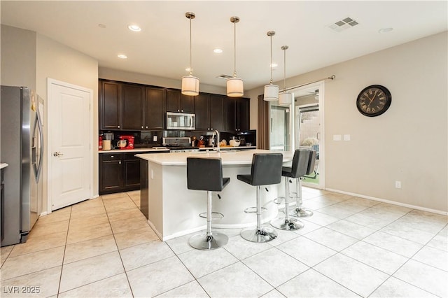 kitchen featuring pendant lighting, light tile patterned floors, stainless steel appliances, dark brown cabinetry, and an island with sink