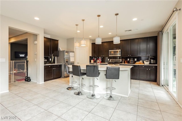 kitchen featuring pendant lighting, light tile patterned floors, a kitchen island with sink, dark brown cabinets, and stainless steel appliances