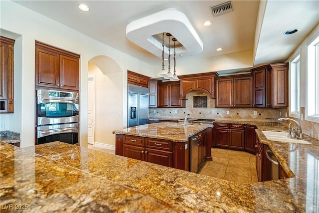 kitchen with sink, a kitchen island with sink, stainless steel appliances, a tray ceiling, and decorative light fixtures