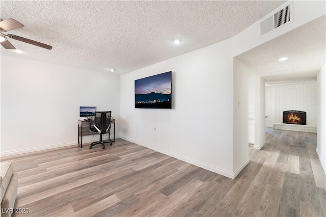 home office featuring ceiling fan, a brick fireplace, a textured ceiling, and light wood-type flooring