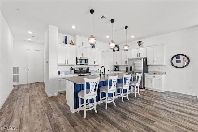 kitchen featuring dark wood-type flooring, white cabinetry, decorative light fixtures, appliances with stainless steel finishes, and an island with sink