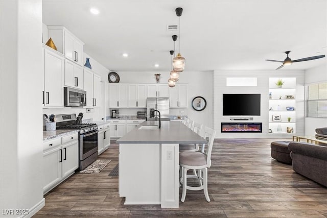 kitchen with dark wood-type flooring, appliances with stainless steel finishes, an island with sink, white cabinets, and decorative light fixtures