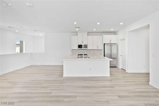 kitchen featuring appliances with stainless steel finishes, white cabinetry, backsplash, a center island with sink, and light hardwood / wood-style flooring