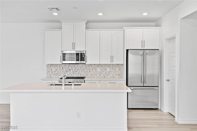 kitchen with a kitchen island with sink, stainless steel appliances, tasteful backsplash, white cabinets, and light wood-type flooring