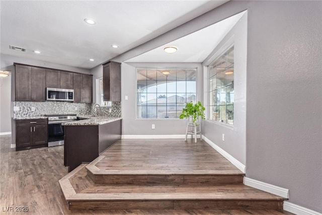 kitchen featuring appliances with stainless steel finishes, decorative backsplash, dark brown cabinetry, light stone countertops, and dark wood-type flooring