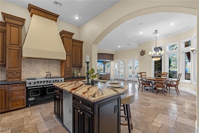 kitchen with custom exhaust hood, range with two ovens, a kitchen island, dark stone counters, and decorative backsplash