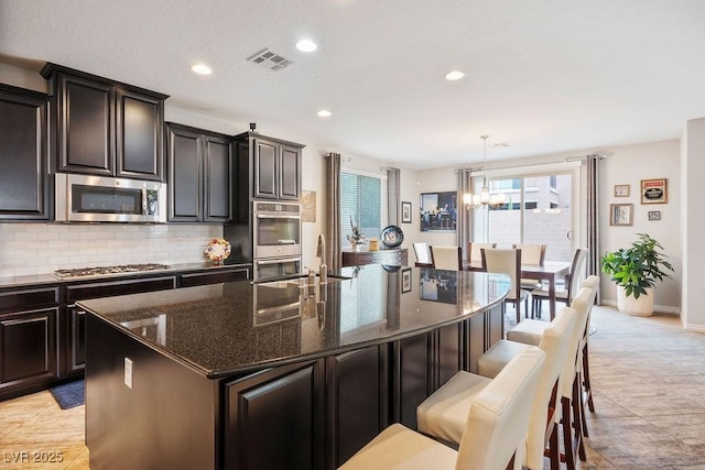 kitchen featuring dark stone counters, an island with sink, pendant lighting, stainless steel appliances, and backsplash