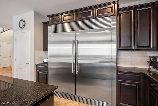 kitchen featuring decorative backsplash, dark stone counters, stainless steel built in fridge, and dark brown cabinetry
