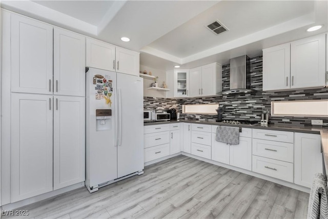 kitchen featuring white cabinets, backsplash, white refrigerator with ice dispenser, a tray ceiling, and wall chimney exhaust hood