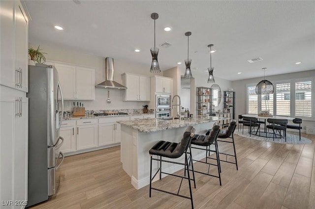 kitchen with white cabinetry, hanging light fixtures, wall chimney range hood, stainless steel appliances, and a center island with sink